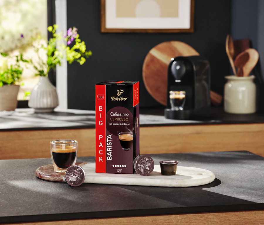 Bir bardak espresso, bir mutfak masasının üzerinde bir paket Tchibo Cafissimo Barista kapsülünün yanında duruyor.