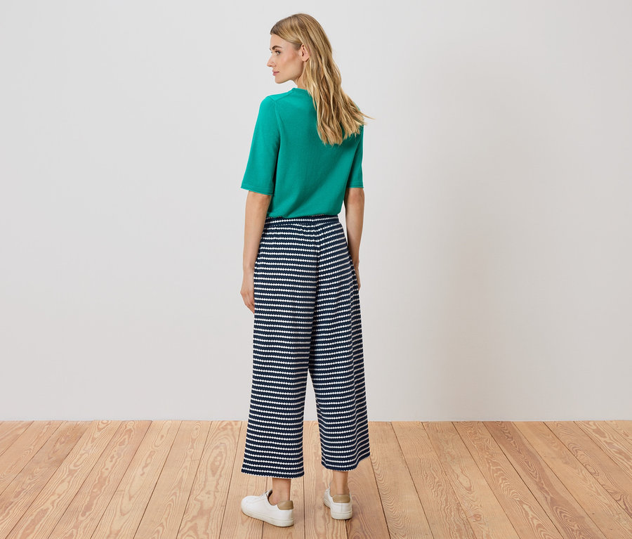 Bir kadın kameraya arkası dönük duruyor, yeşil bir üst ve lacivert-beyaz çizgili culottes pantolon giyiyor.