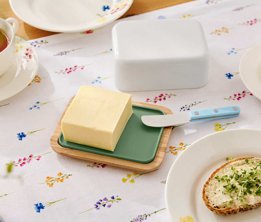 Çiçekli bir masa üzerinde, yeşil ek parçalı ahşap bir tabakta tereyağı, tereyağı bıçağı, tereyağlık, bir fincan çay ve tereyağlı bir sandviç bulunan bir tabak bulunmaktadır.