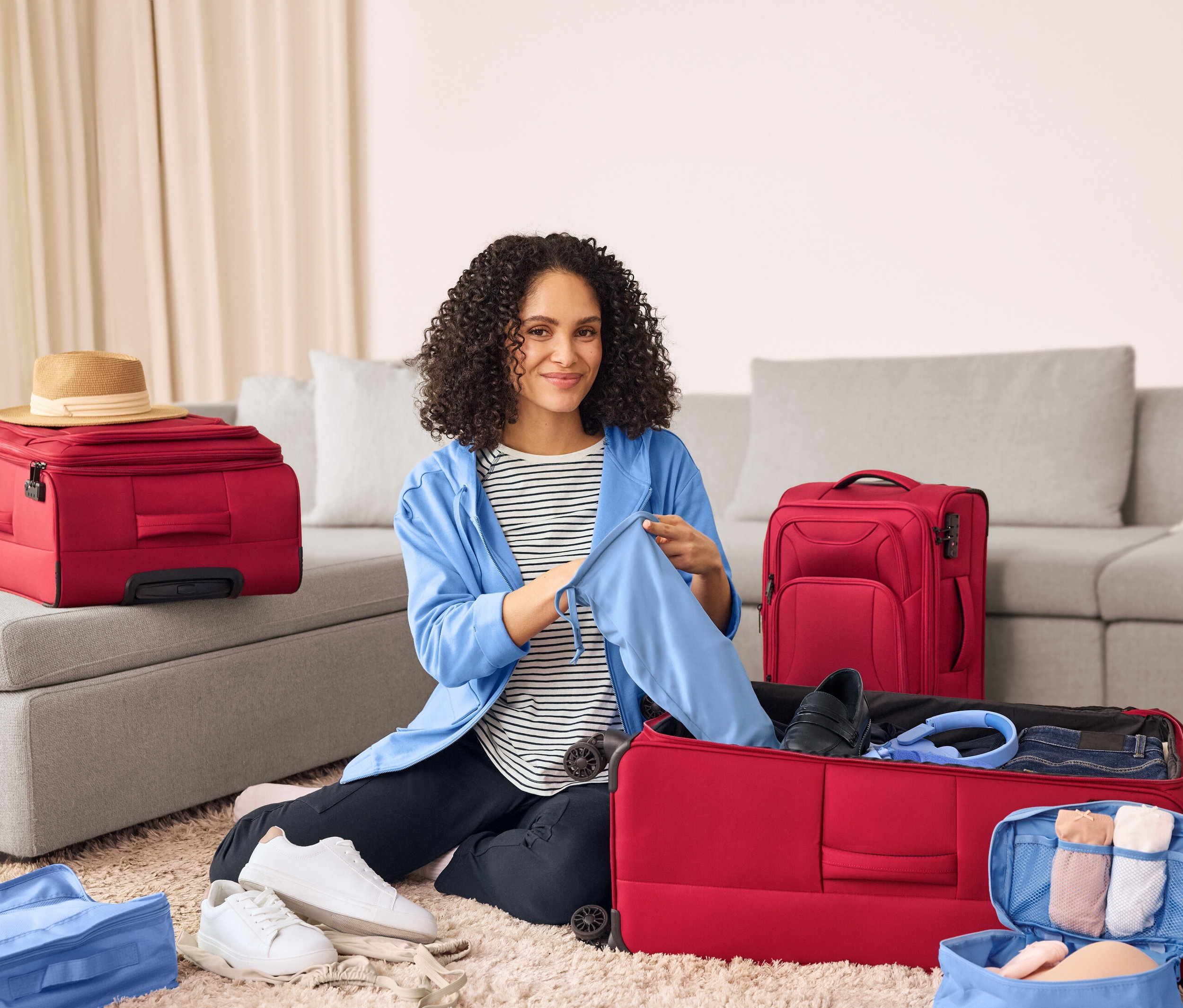 Bir kadın yerde oturuyor ve koyu mavi kargo cepli eşofman altını kırmızı bir valize koyuyor. Yanında deri spor ayakkabılar var.