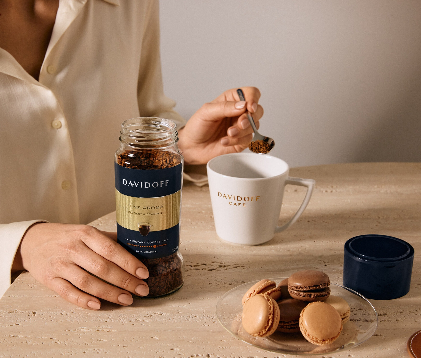 Kadın bir kaşıkla DAVIDOFF CAFÉ Fine Aroma çözünebilir kahveyi fincana döküyor. Tabakta makaronlar var.