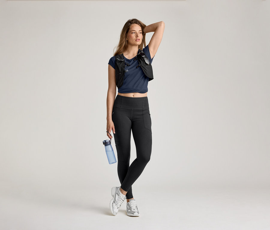 Kadın, elini başına koymuş su şişesiyle beraber outdoor tayt, mavi tişört ve koşu yeleğiyle poz veriyor.