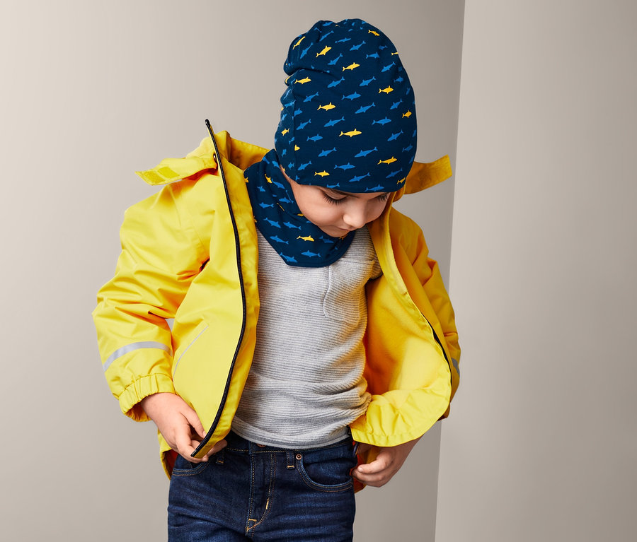 Sarı ceketli bir çocuk ceketinin fermuarını ayarlıyor, köpekbalığı desenli bere ve boyunluk takıyor.