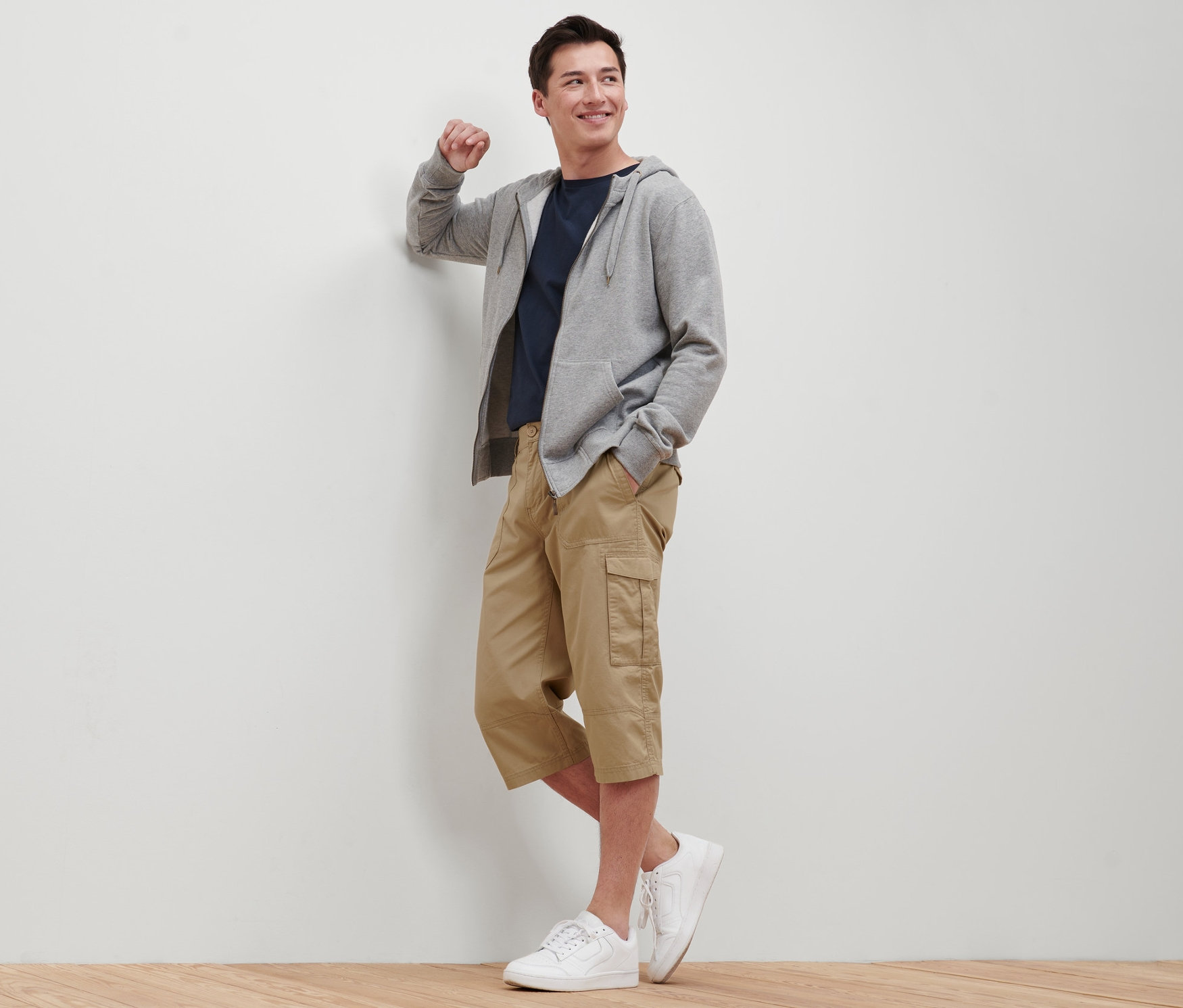 Duvara yaslanmış bir adam bej renkli 3/4 boy kargo bermuda şort, tişört, kapüşonlu sweatshirt ve spor ayakkabı giyiyor.