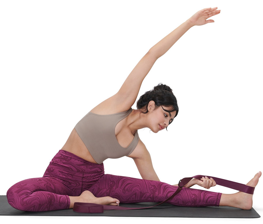 Kadın yoga matında oturuyor ve bacağını esnetmek için mor 2'si 1 Arada Yoga Kemeri kullanıyor.