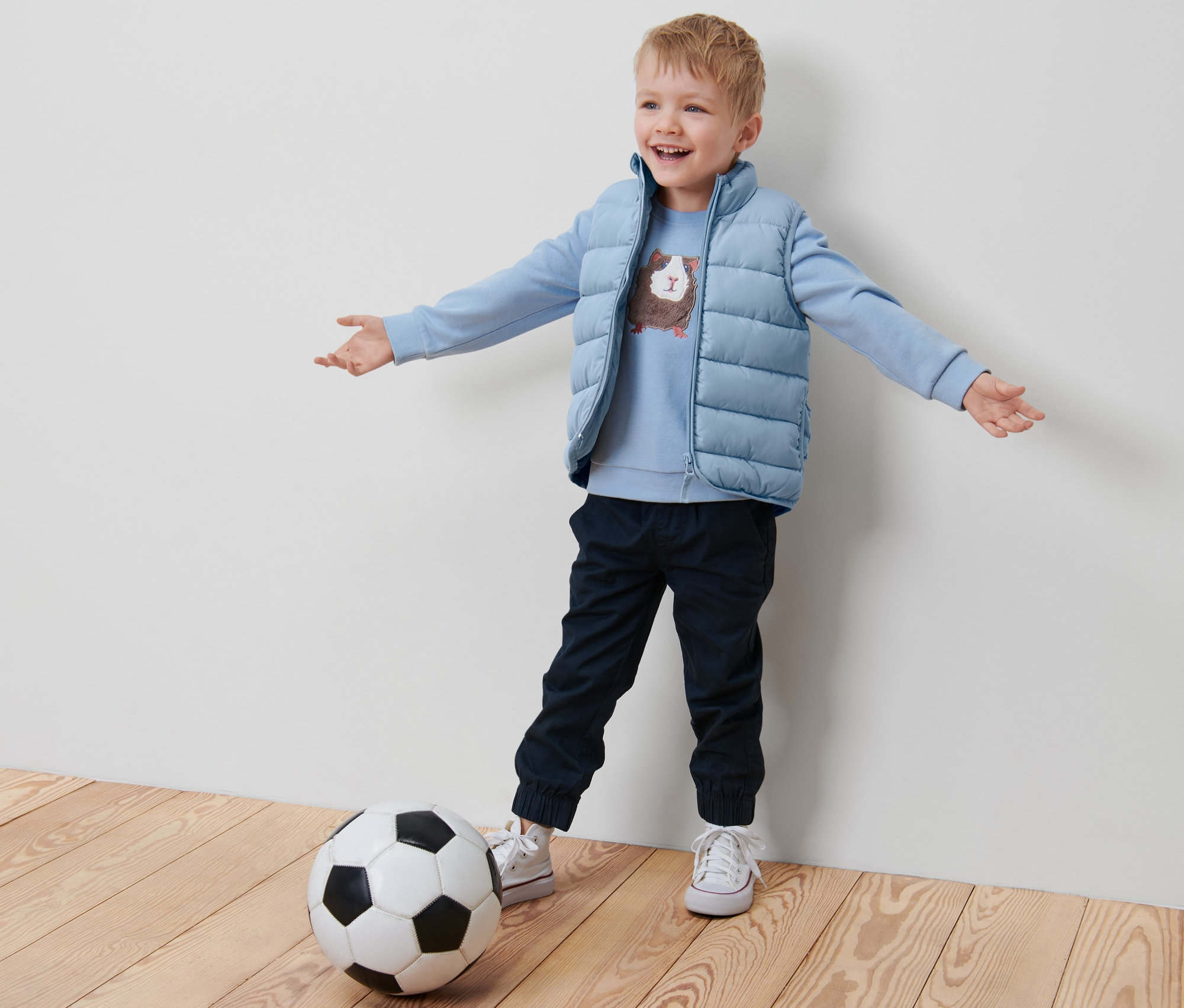 Bir çocuk futbol topunun yanında kolları açık duruyor, üzerinde mavi Küçük Çocuk Kapitone Yelek var.