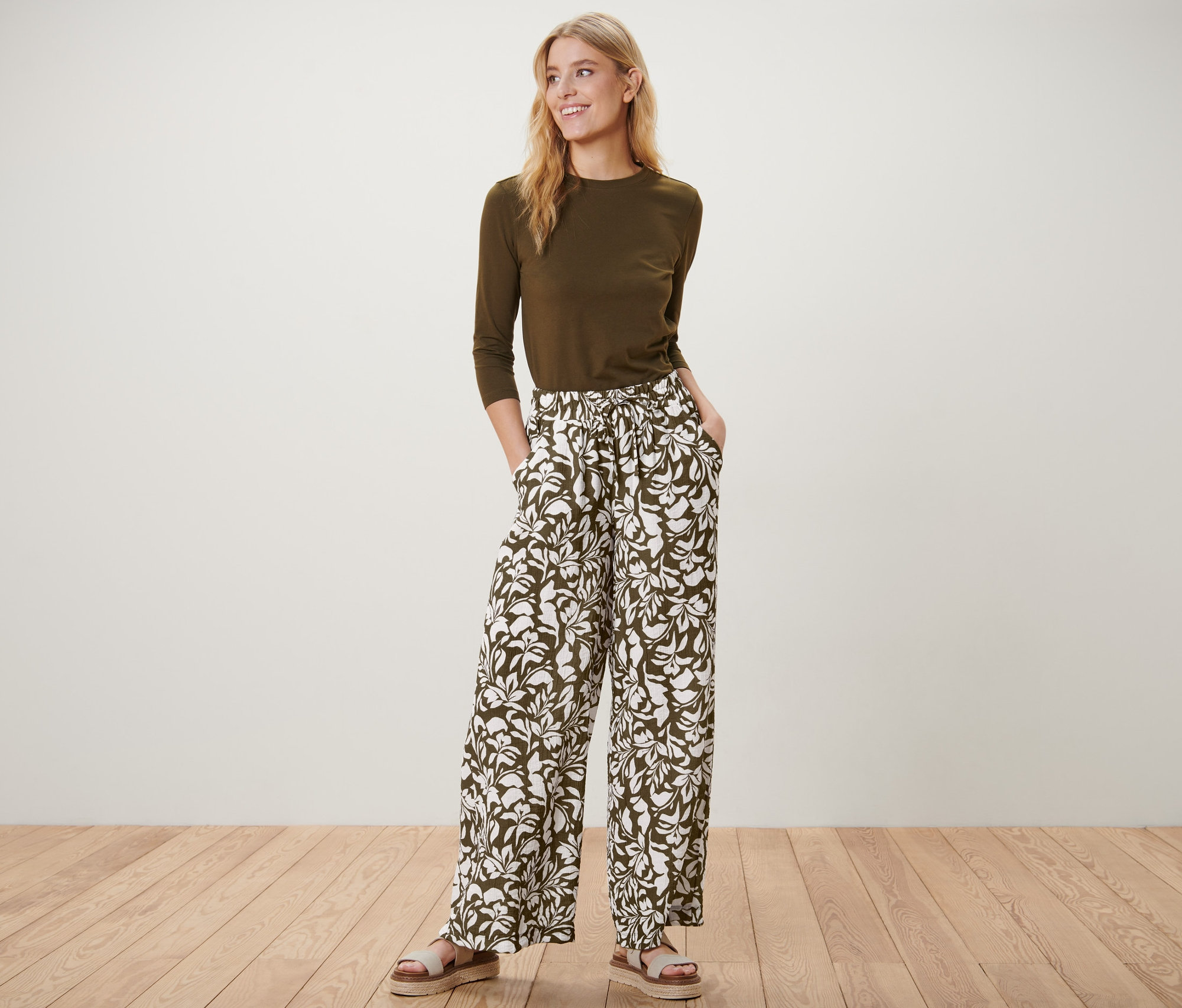 Kadın baskılı eşofman pantolonu, zeytin yeşili üstü ve sandaletleriyle ayakta poz veriyor.