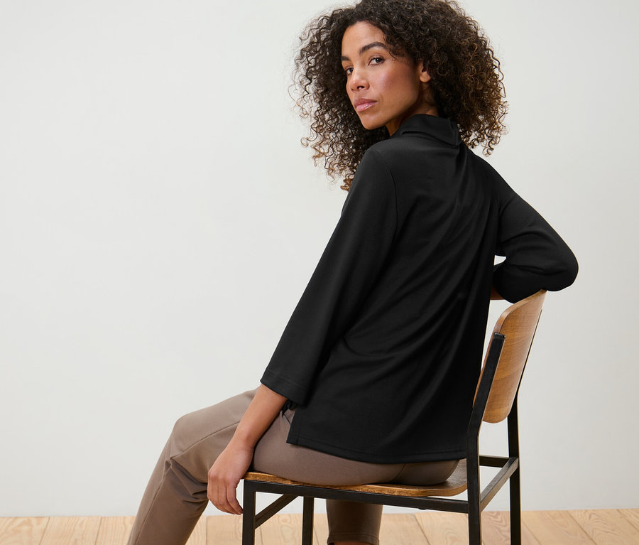 Kadın sandalyede oturuyor ve siyah, 3/4 kollu bir sweatshirt giyiyor.