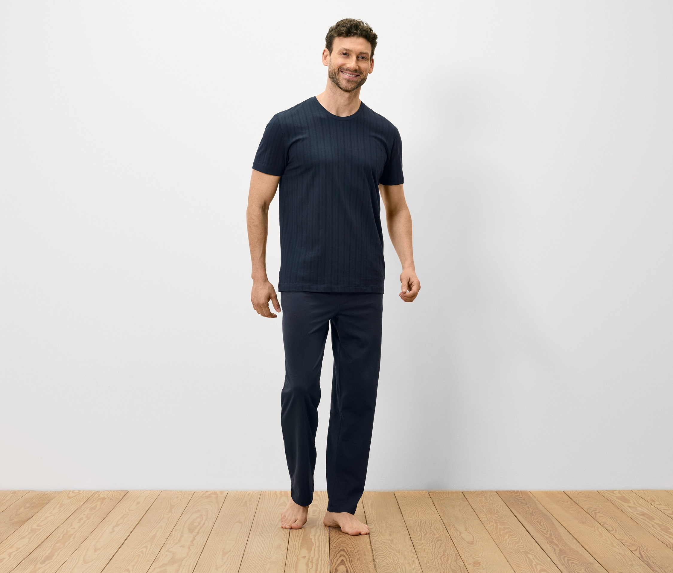 Adam ahşap zeminde çıplak ayakla duruyor ve koyu mavi bir pijama giyiyor.