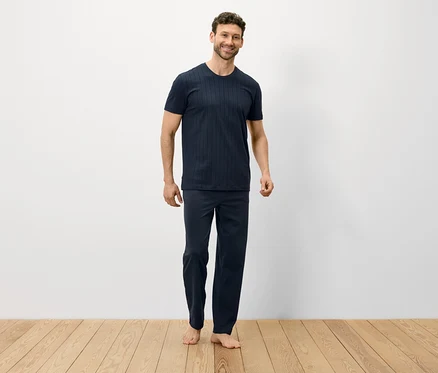 Adam ahşap zeminde çıplak ayakla duruyor ve koyu mavi bir pijama giyiyor.