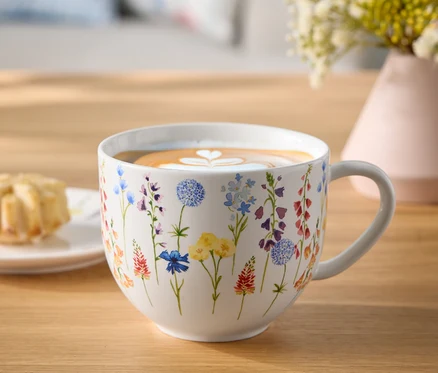 Çiçekli fincan, kahve ve latte sanatıyla ahşap bir masanın üzerinde, yanında bir tabak hamur işi ve çiçek vazosu bulunuyor.