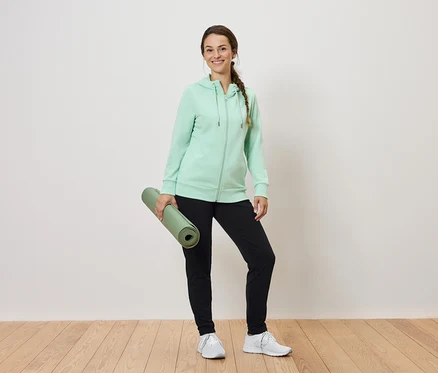Bir kadın rulo halinde yeşil bir yoga matı tutuyor, üzerinde nane yeşili bir kapüşonlu sweatshirt ve siyah eşofman altı var.