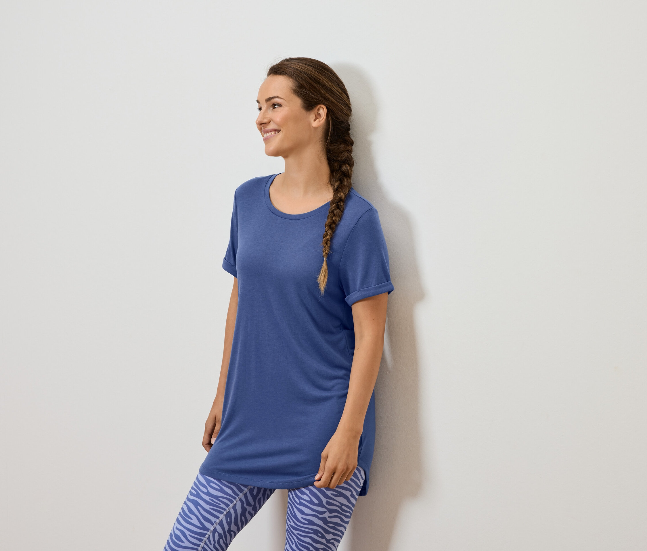 Kahverengi örgülü bir kadın beyaz bir duvarın önünde duruyor, mavi bir tişört ve hayvan desenli mavi tayt giyiyor.