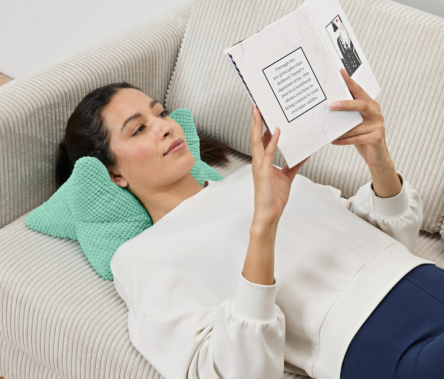 Bir kadın kanepede uzanmış kitap okuyor, başı bir okuma yastığı üzerinde duruyor.