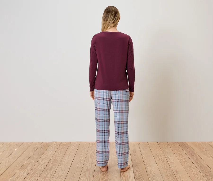 Bir kadın kameraya arkası dönük duruyor, bordo bir kazak ve mavi ekose pijama altı giyiyor.