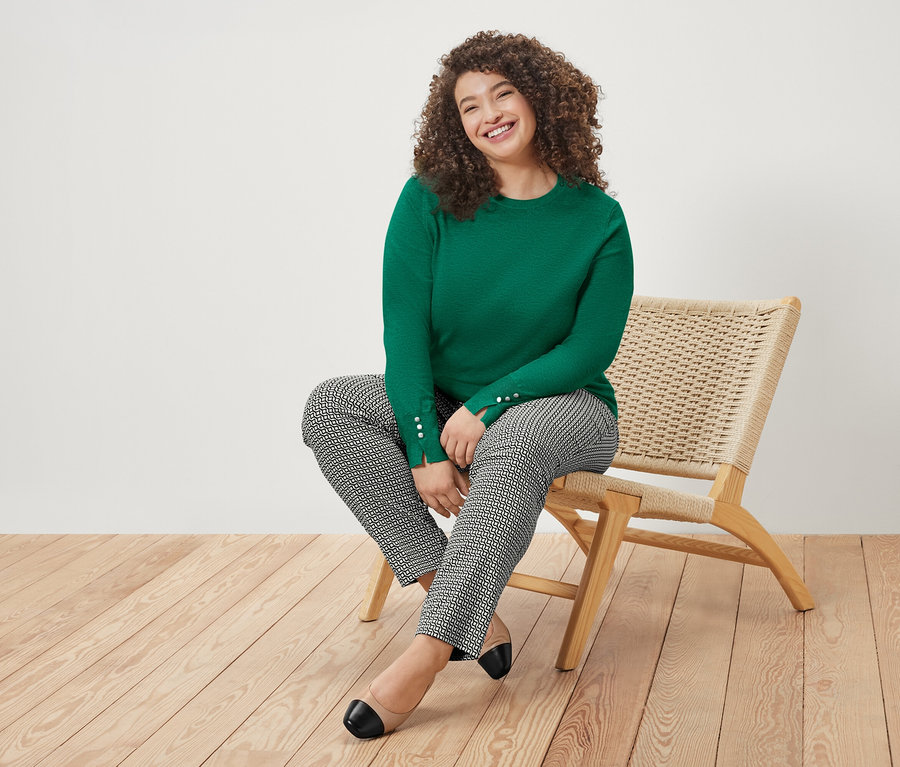 Kıvırcık saçlı bir kadın sandalyede oturuyor, yeşil bir kazak ve desenli pantolon giyiyor.