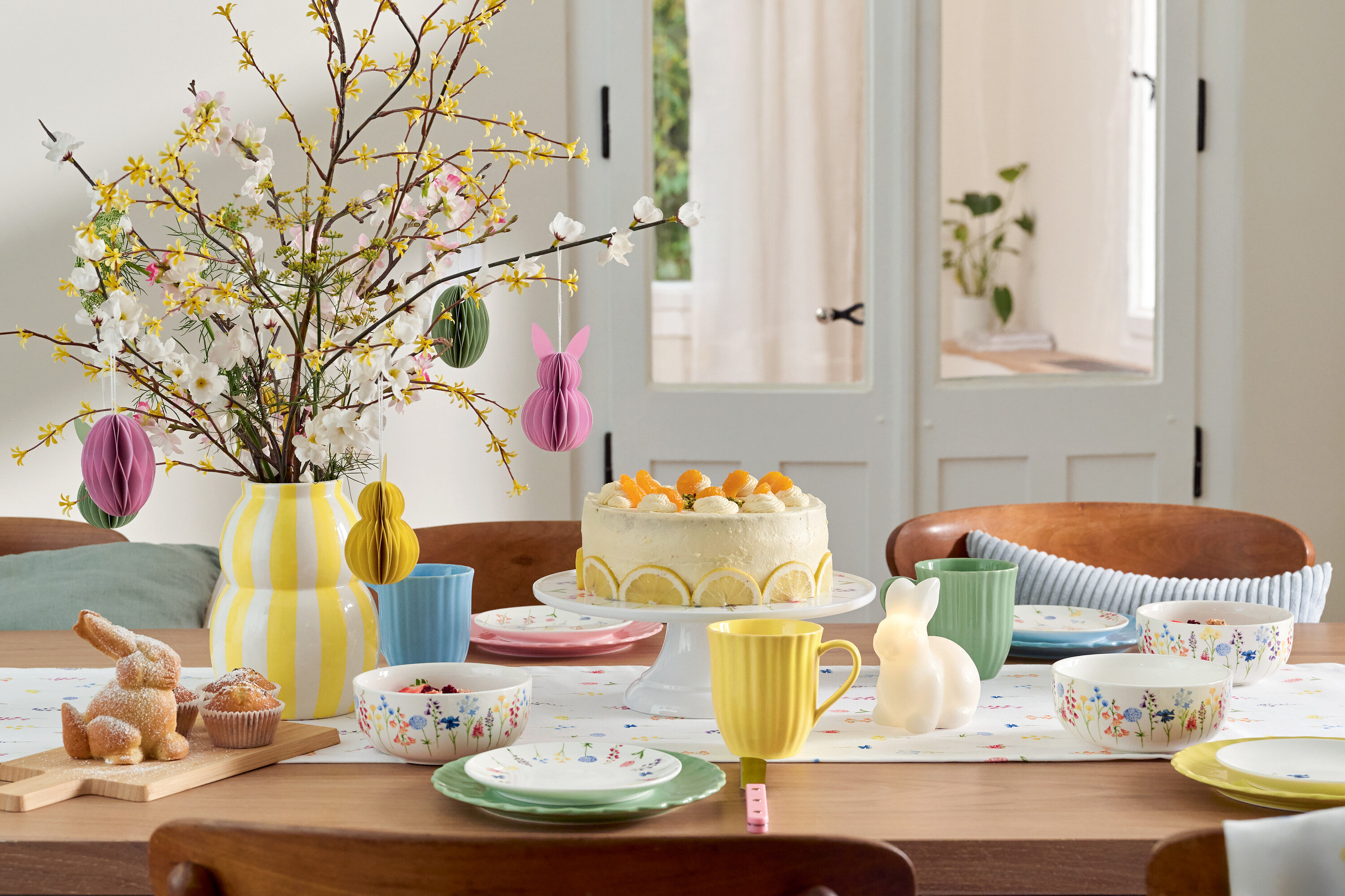 Paskalya dekorasyonları, pasta, sofra takımı ve çiçek vazosu olan yemek masası.