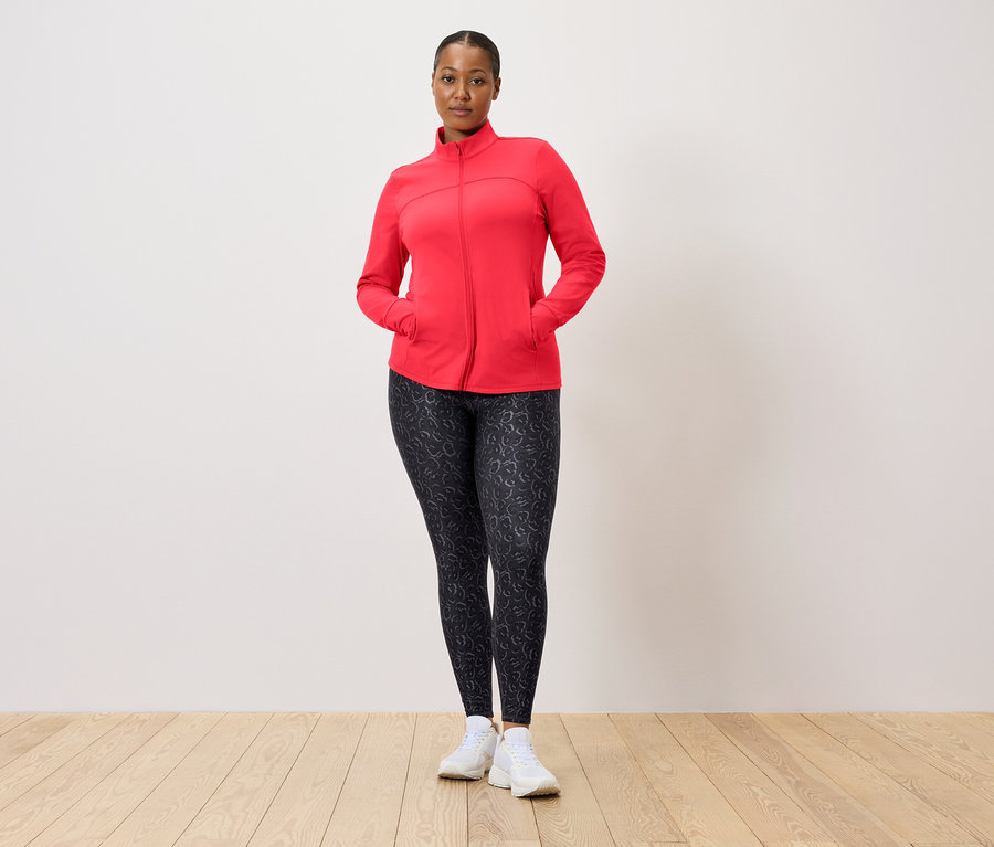 Kırmızı ceketli ve desenli taytlı bir kadın duvar önünde poz veriyor.