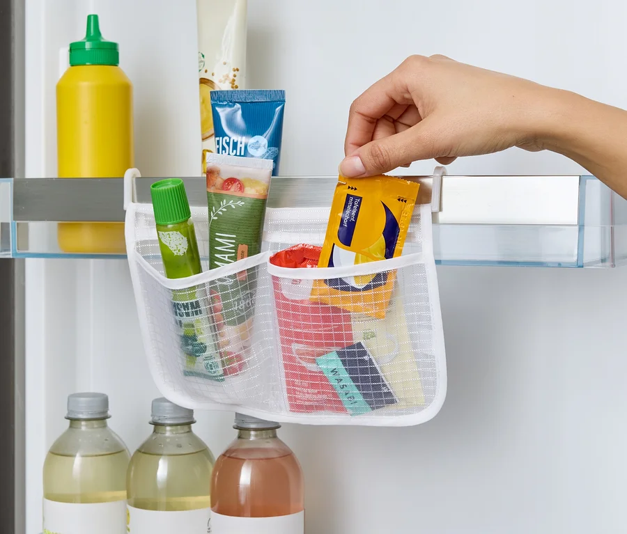 Bir el, buzdolabı rafına asılı beyaz file organizerin içine sarı bir paket yerleştiriyor. İçinde tüpler, soslar ve wasabi var.