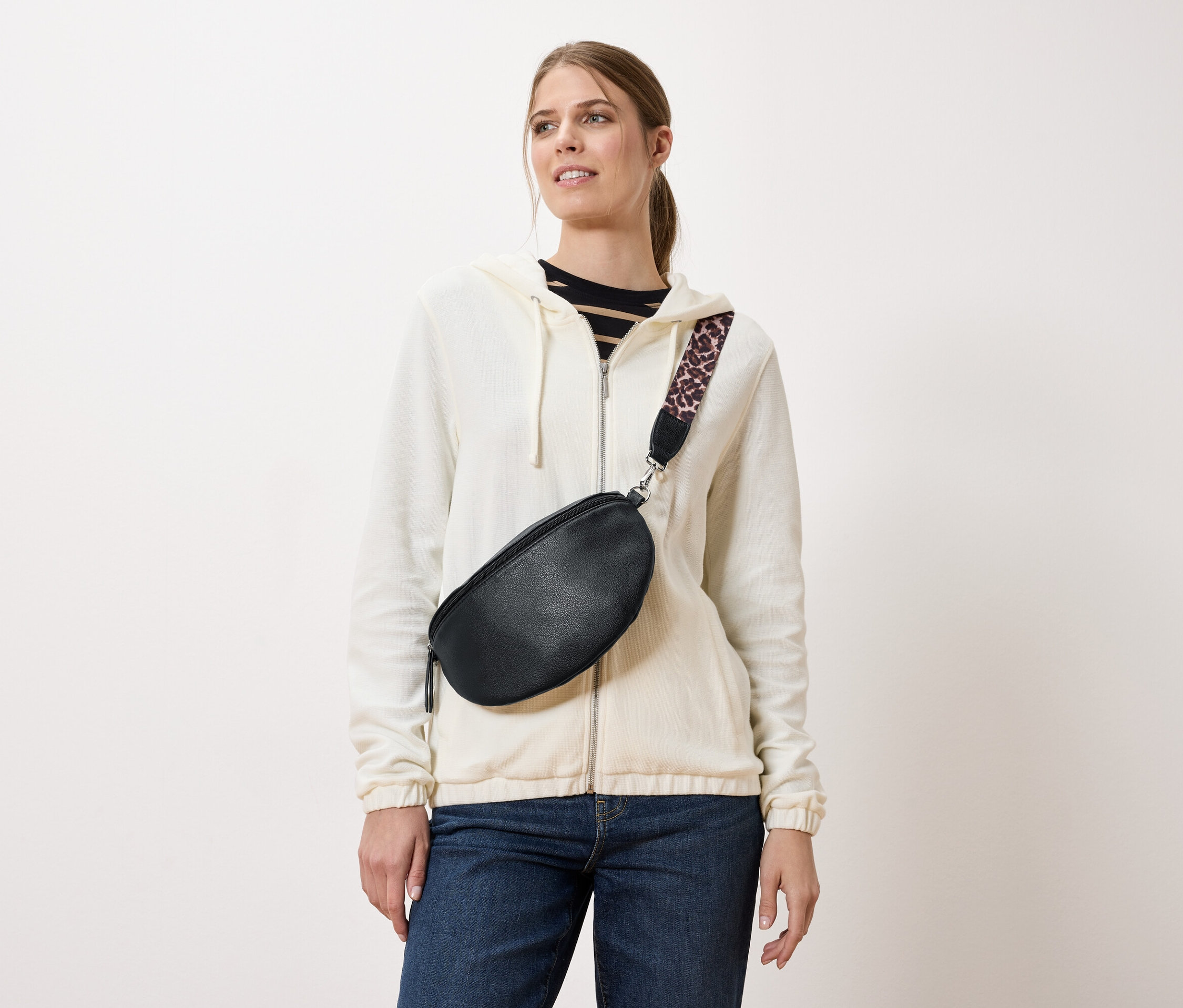 Bir kadın, krem rengi bir ceket ve kot pantolon üzerine takılmış leopar desenli askılı siyah bir bel çantası takıyor.