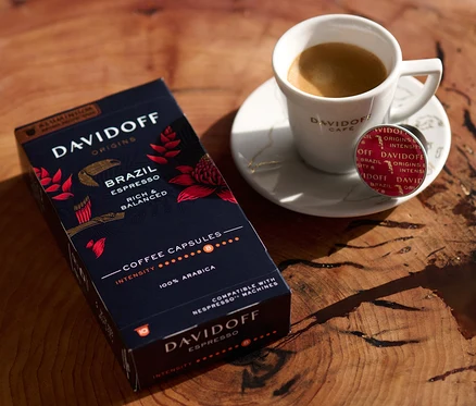 Ahşap bir masanın üzerinde bir paket DAVIDOFF CAFÉ ORIGINS Brazil Espresso, bir fincan espresso ve bir kapsül var.