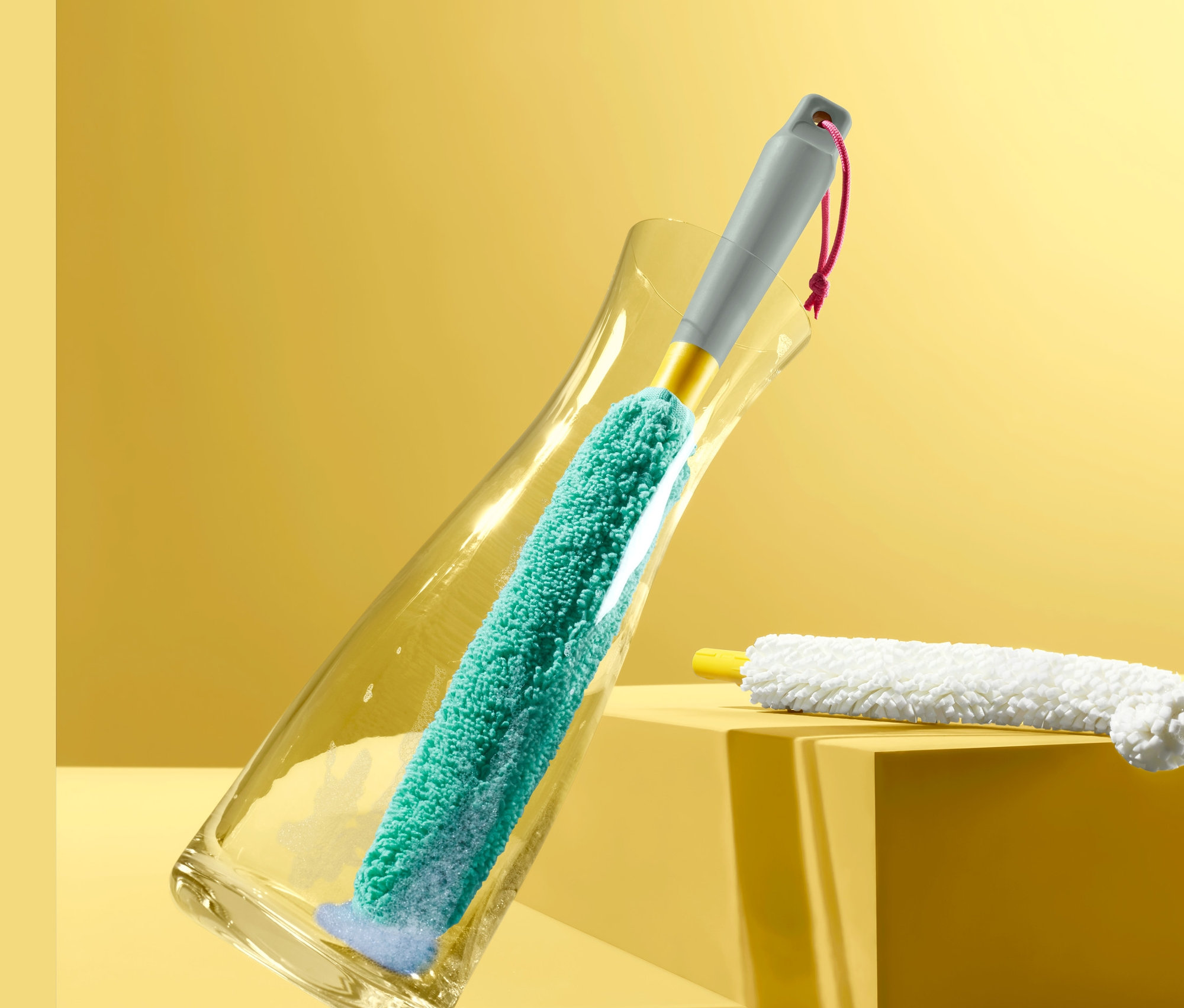 Şeffaf bir vazoda yeşil renkli 2'si 1 Arada: Şişe ve Cam Fırçası var. İkinci bir fırça vazonun yanında duruyor.