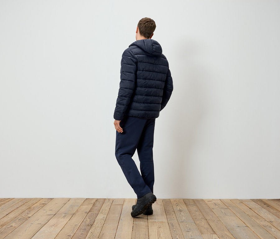 Arkası kameraya dönük bir adam ahşap zeminde duruyor, lacivert kapüşonlu bir ceket ve pantolon giyiyor.