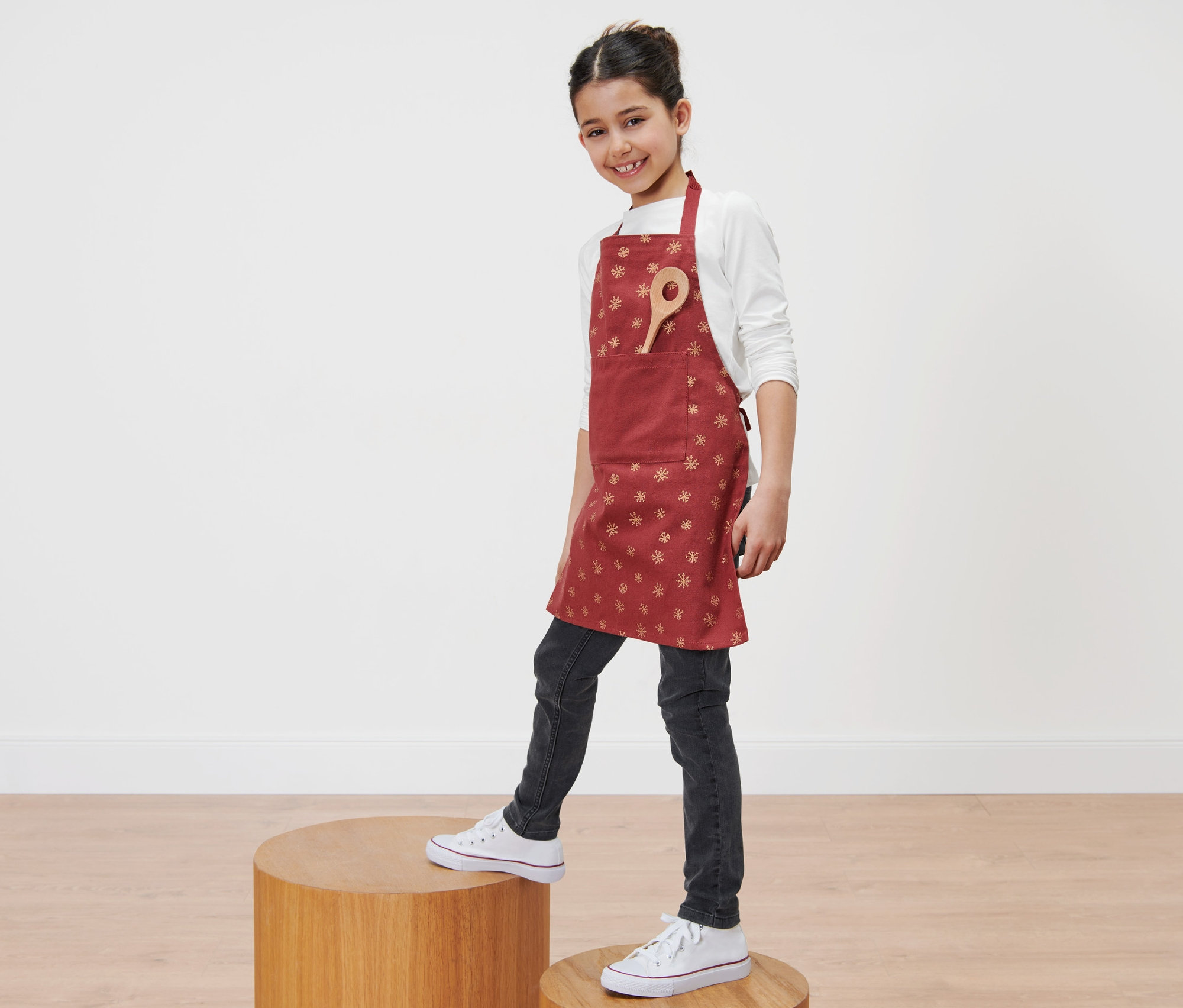 Kız tahta bir platformda duruyor ve kar tanesi desenli kırmızı bir Çocuk Önlüğü giyiyor.
