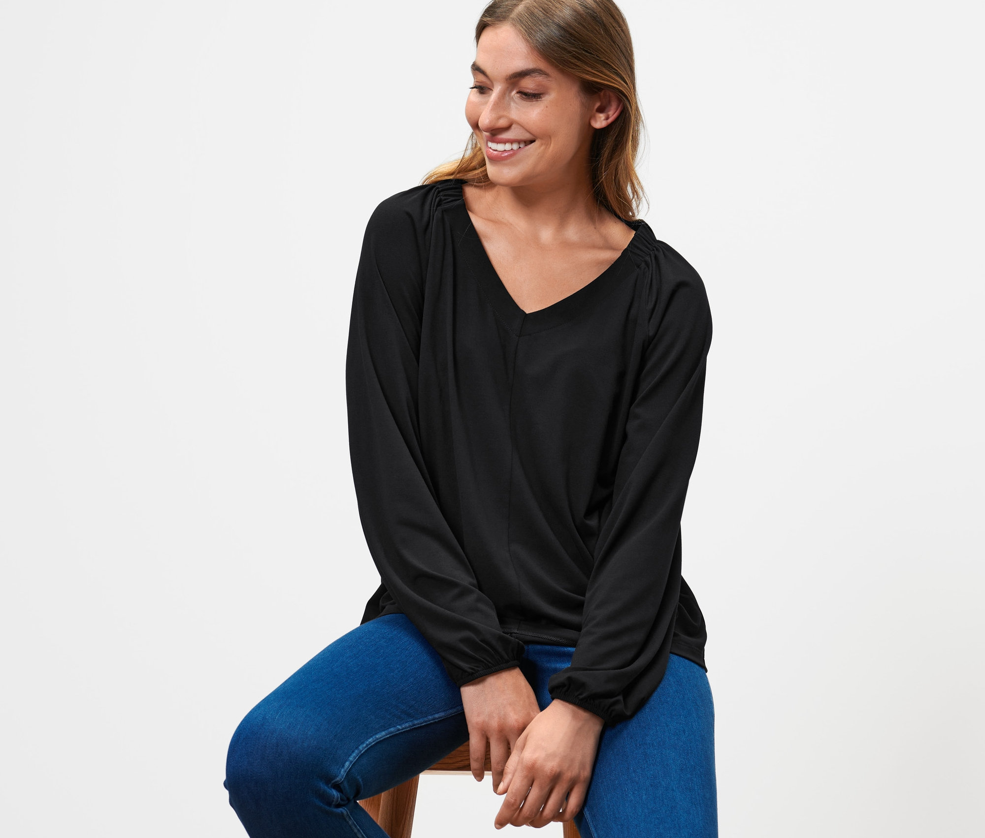 Kadın bir taburede oturuyor, üzerinde siyah büzgülü bluz ve mavi kot pantolon var.