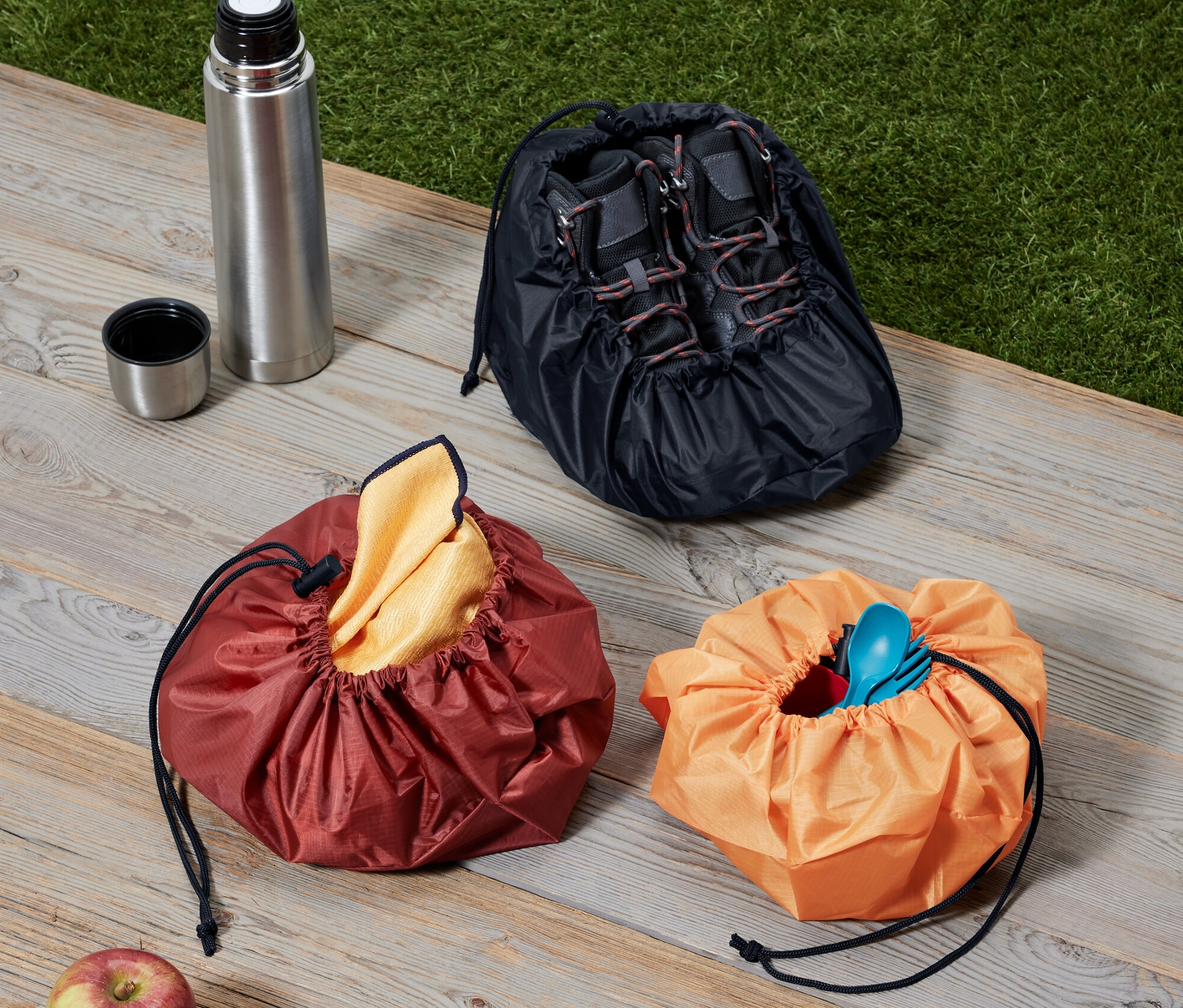 Tahta bir yüzeyde üç adet çanta, bir termos ve bir elma duruyor.