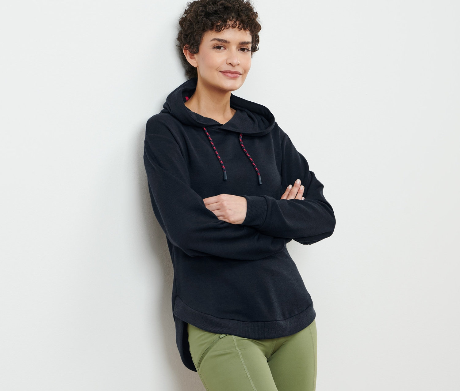 Kahverengi kıvırcık saçlı kadın, siyah kapüşonlu sweatshirt ve yeşil pantolonla poz veriyor.