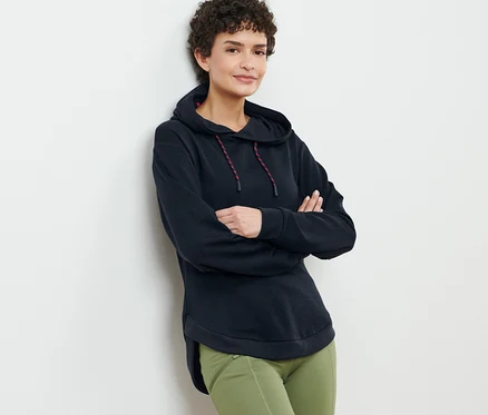 Kahverengi kıvırcık saçlı kadın, siyah kapüşonlu sweatshirt ve yeşil pantolonla poz veriyor.