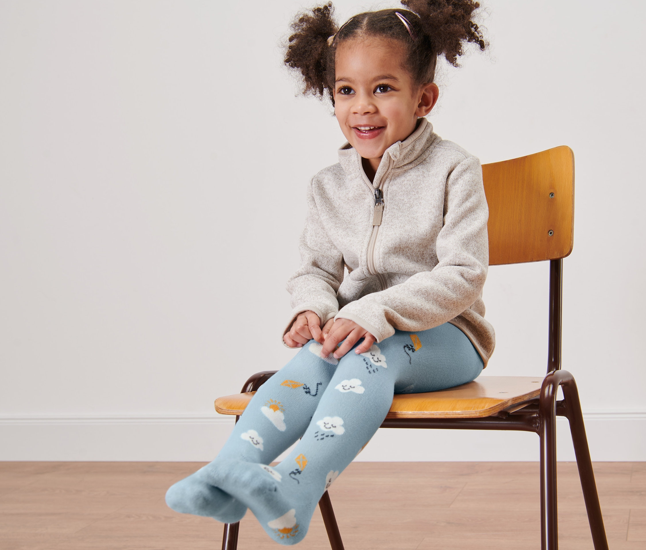 Gülümseyen küçük kız bir sandalyede oturuyor ve bulut desenli mavi külotlu çorap giyiyor.