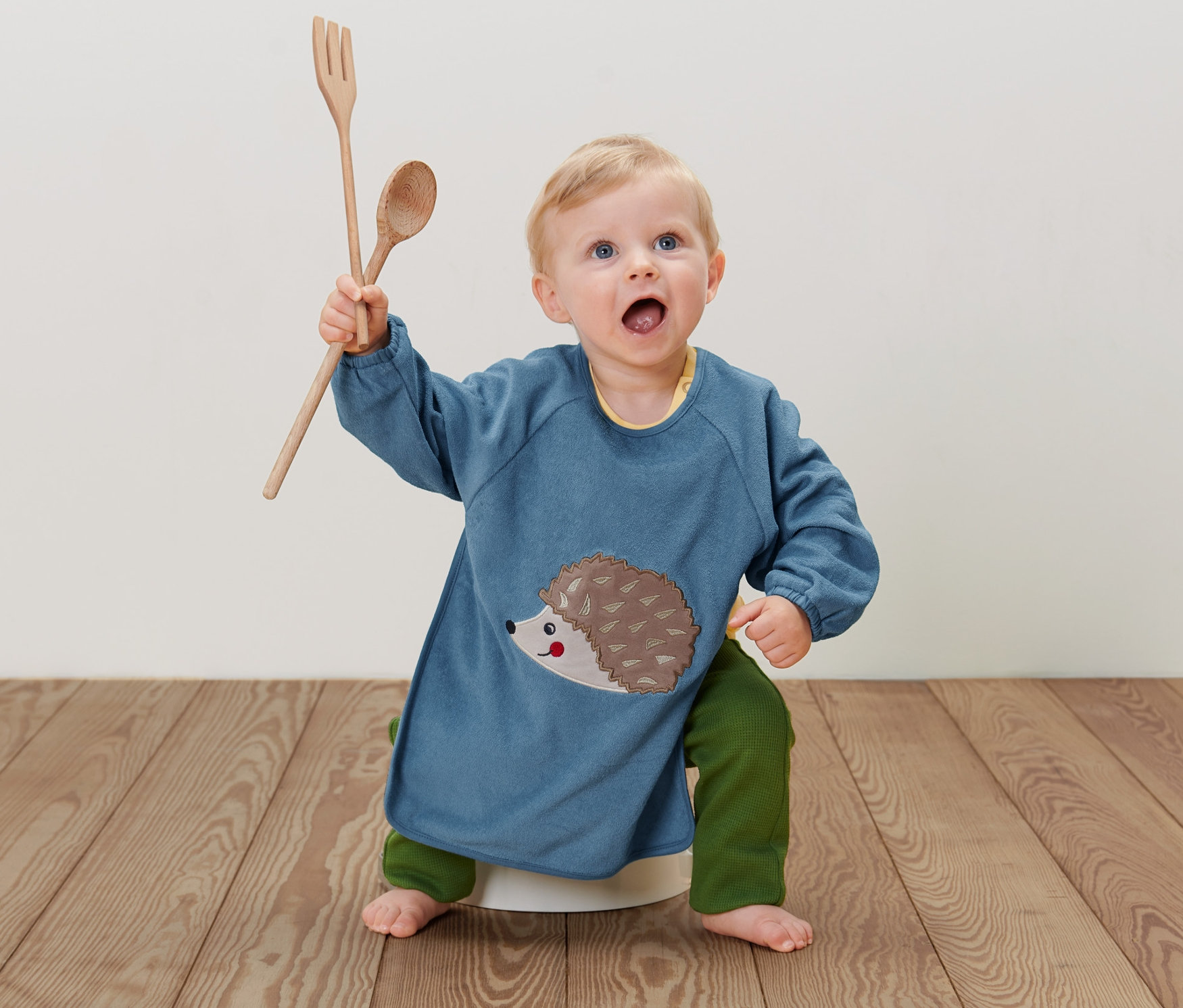 Bebek yerde oturuyor, üzerinde kirpili mavi 2 Adet Kollu Bebek Mama Önlüğü var, elinde tahta kaşık ve çatal tutuyor.