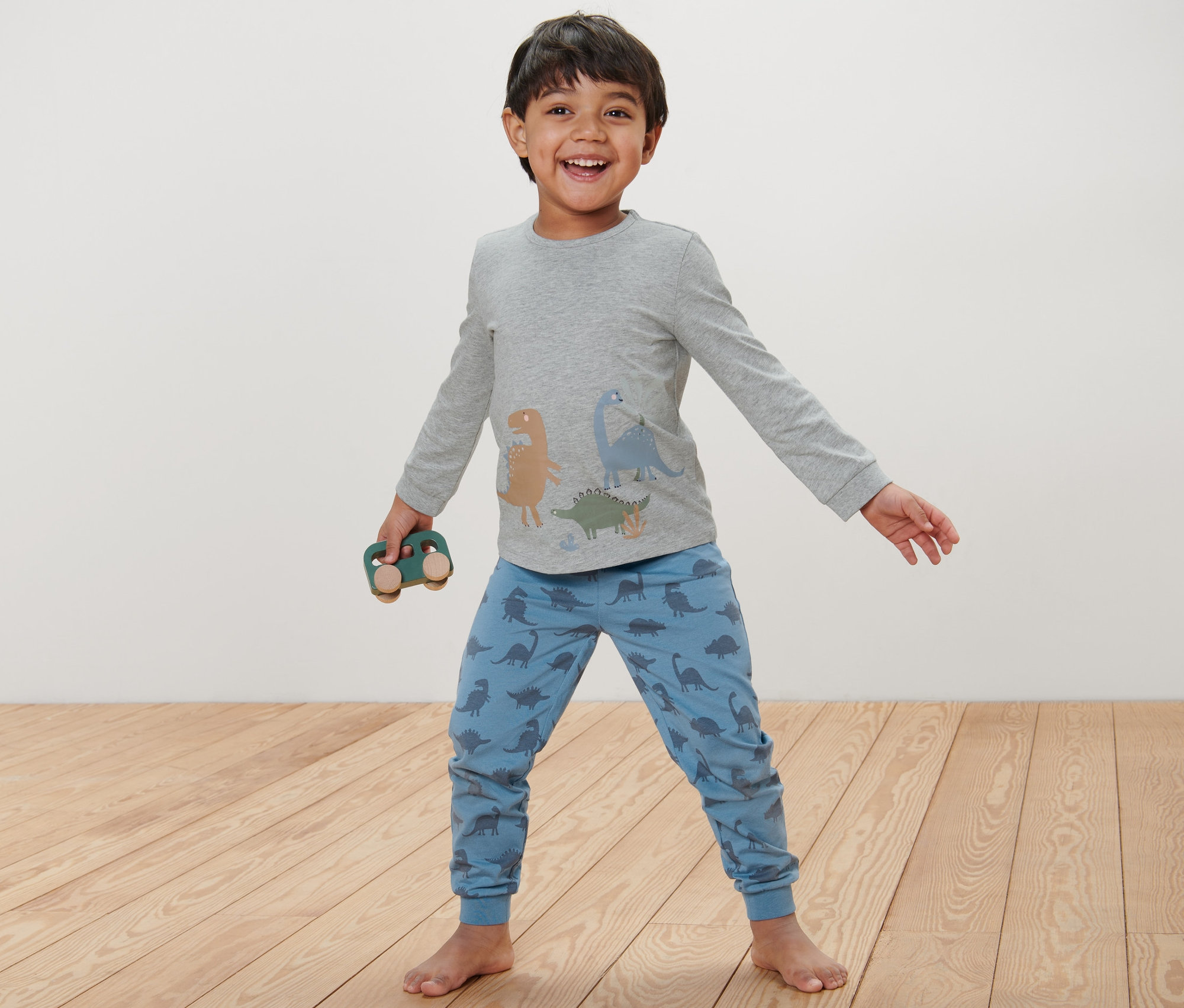 Dinozor desenli mavi çocuk pijaması giyen bir çocuk, tahta oyuncak araba tutuyor.