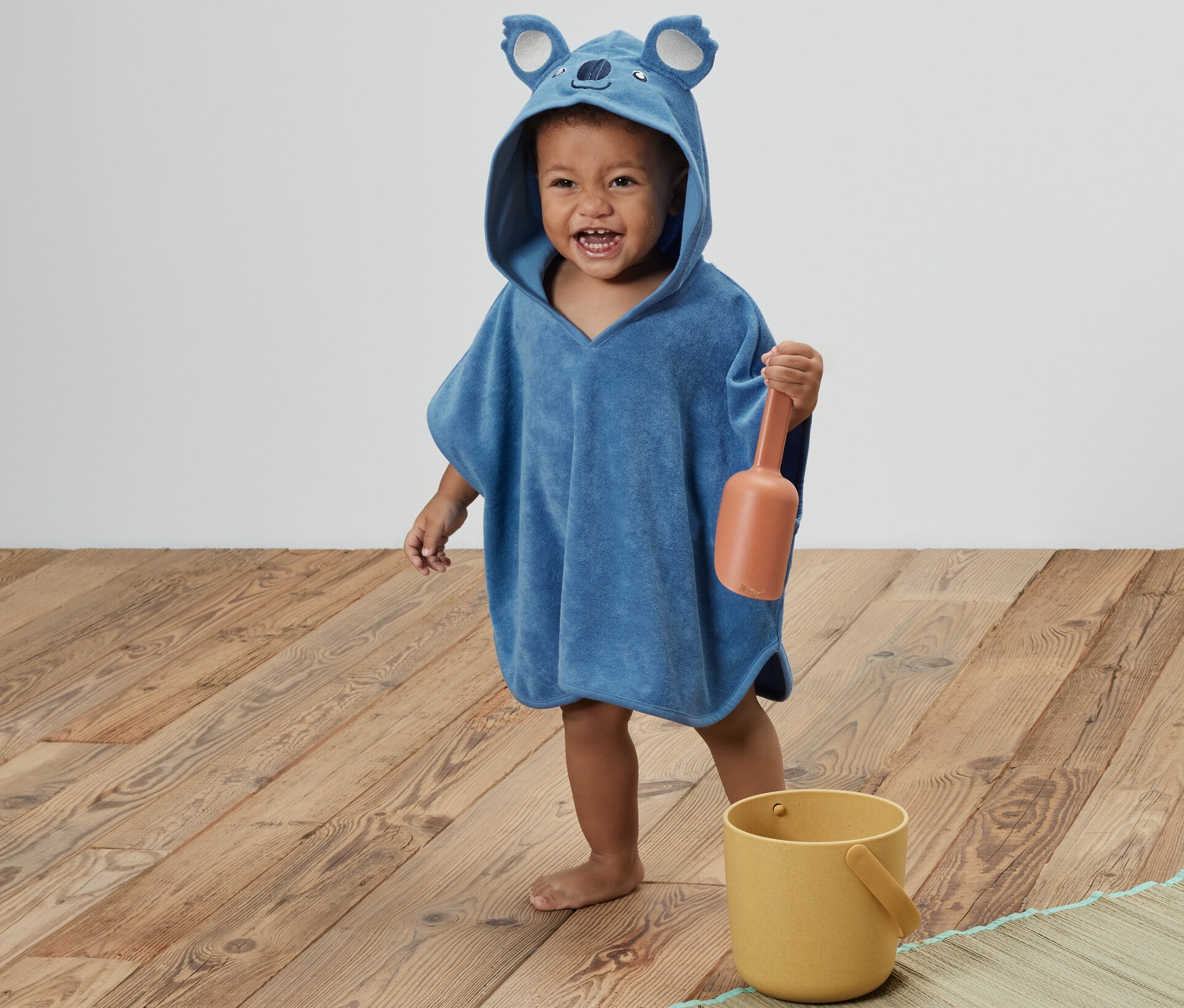 Mavi, koala kapüşonlu çocuk havlu panço giyen bir çocuk, turuncu bir kürek tutuyor. Yanında sarı bir kova duruyor.