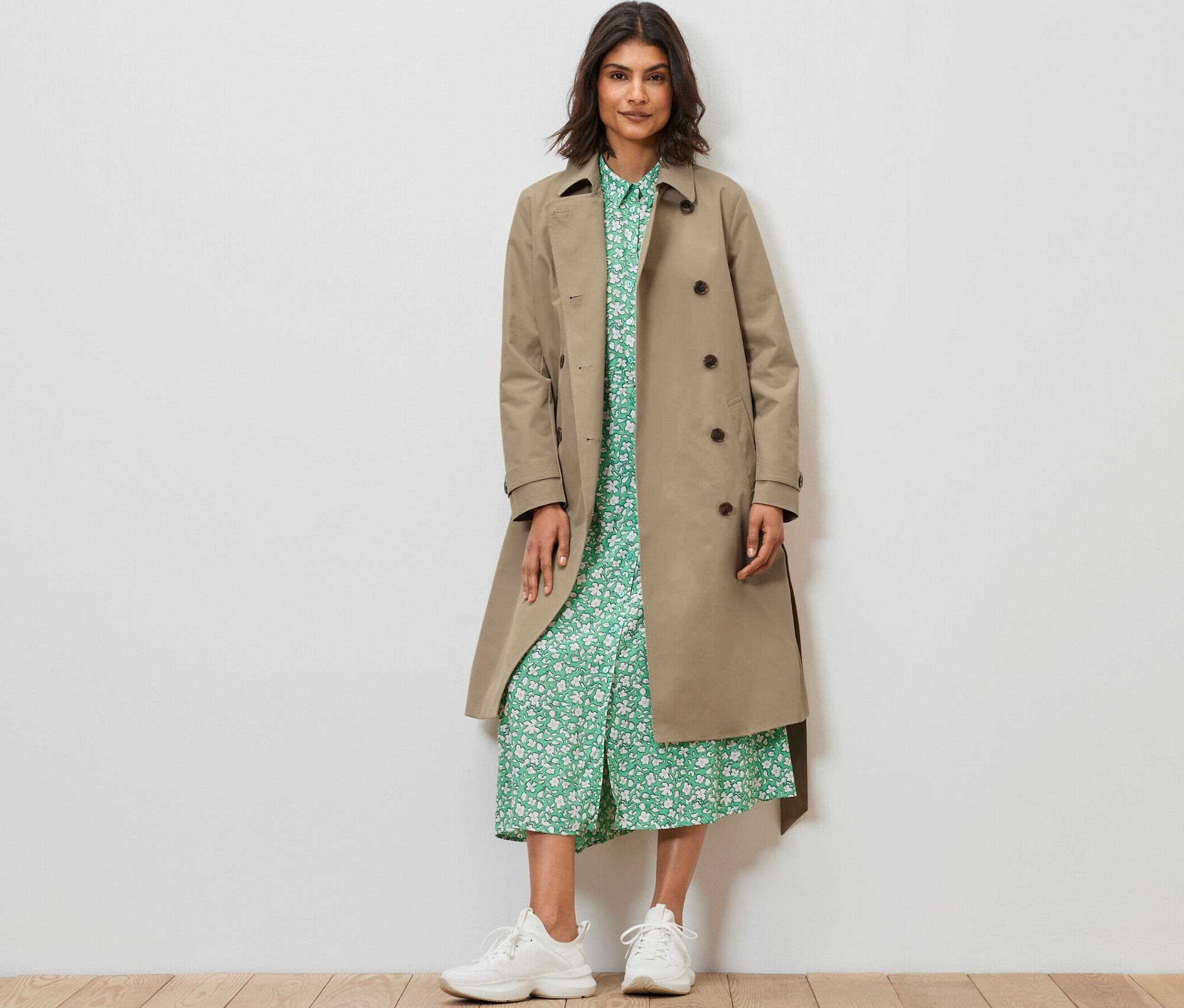 Kadın ayakta duruyor ve Kum Beji renginde bir trençkot giyiyor.