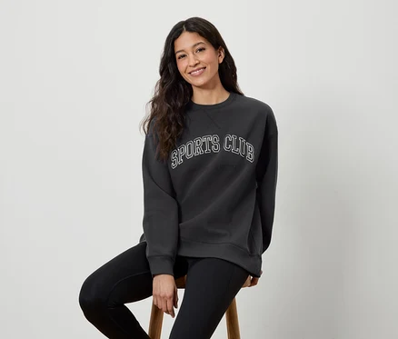 Bir kadın taburede oturuyor, üzerinde SPORTS CLUB yazılı koyu gri bir sweatshirt ve siyah tayt var.