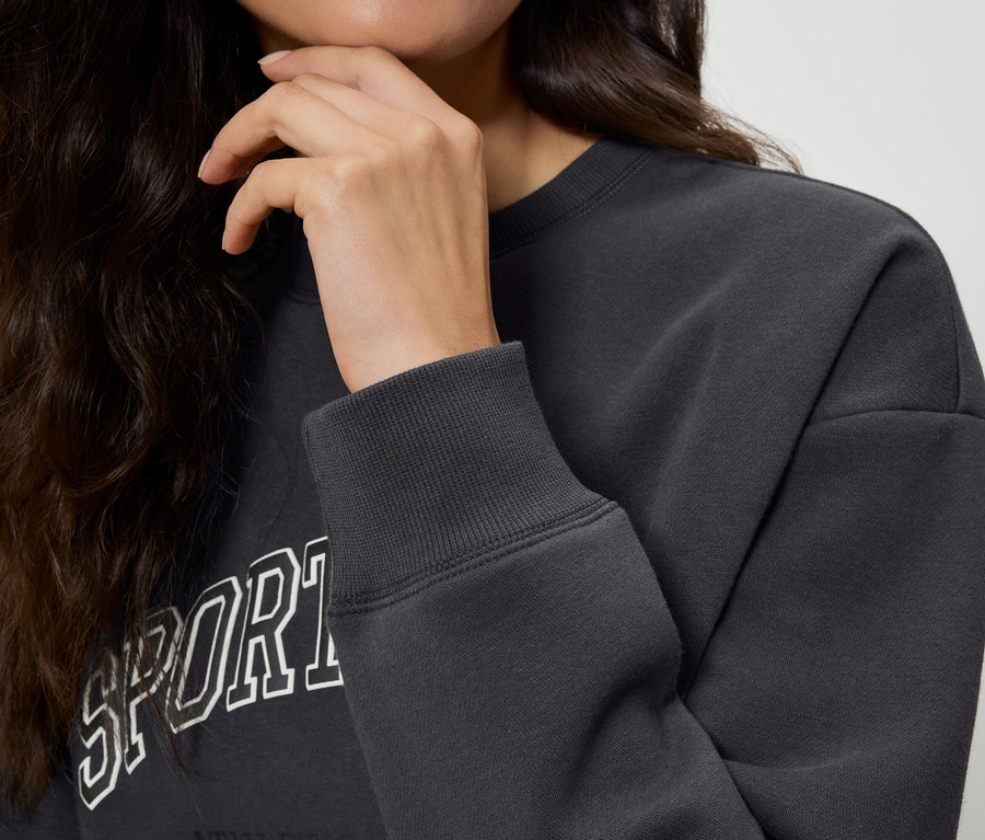 Çenesiyle temas halinde, dalgalı koyu renk saçlı bir kadının yakın çekimi. Üzerinde Sport yazılı koyu gri bir sweatshirt var. Sweatshirt'ün kolunun yakından çekilmiş hali.