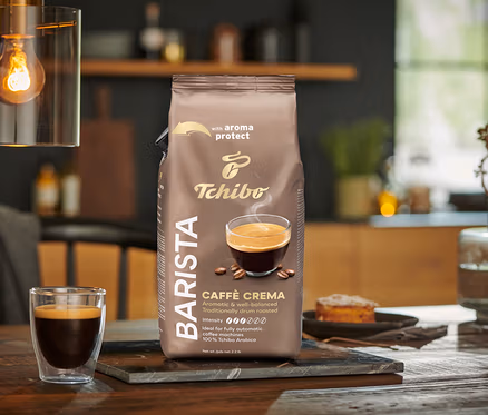 Bir paket BARISTA Caffè Crema - 1x 1 kg Çekirdek Kahve, bir bardak kahvenin yanında masanın üzerinde duruyor.