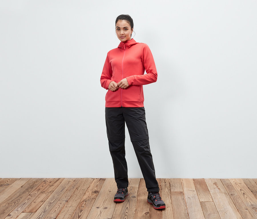 Bir kadın, ahşap zeminde mercan rengi kapüşonlu bir ceket, siyah pantolon ve ayakkabılarla poz veriyor.