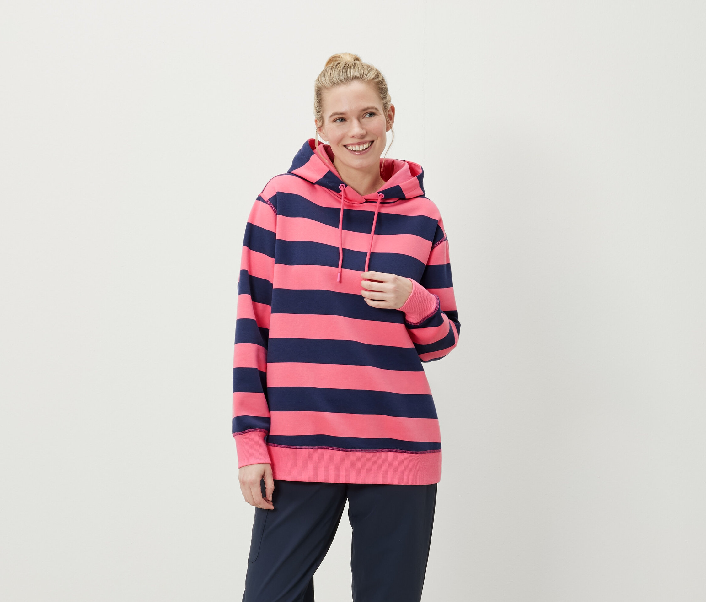 Sarışın saçlı, gülümseyen bir kadın, pembe ve lacivert çizgili kapüşonlu bir sweatshirt ve lacivert pantolon giyiyor.