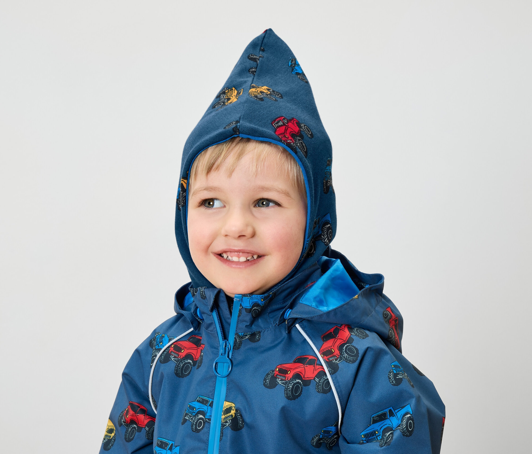 Sırıtan bir çocuk, canavar kamyon baskılı mavi bir ceket ve kapüşon giyiyor.