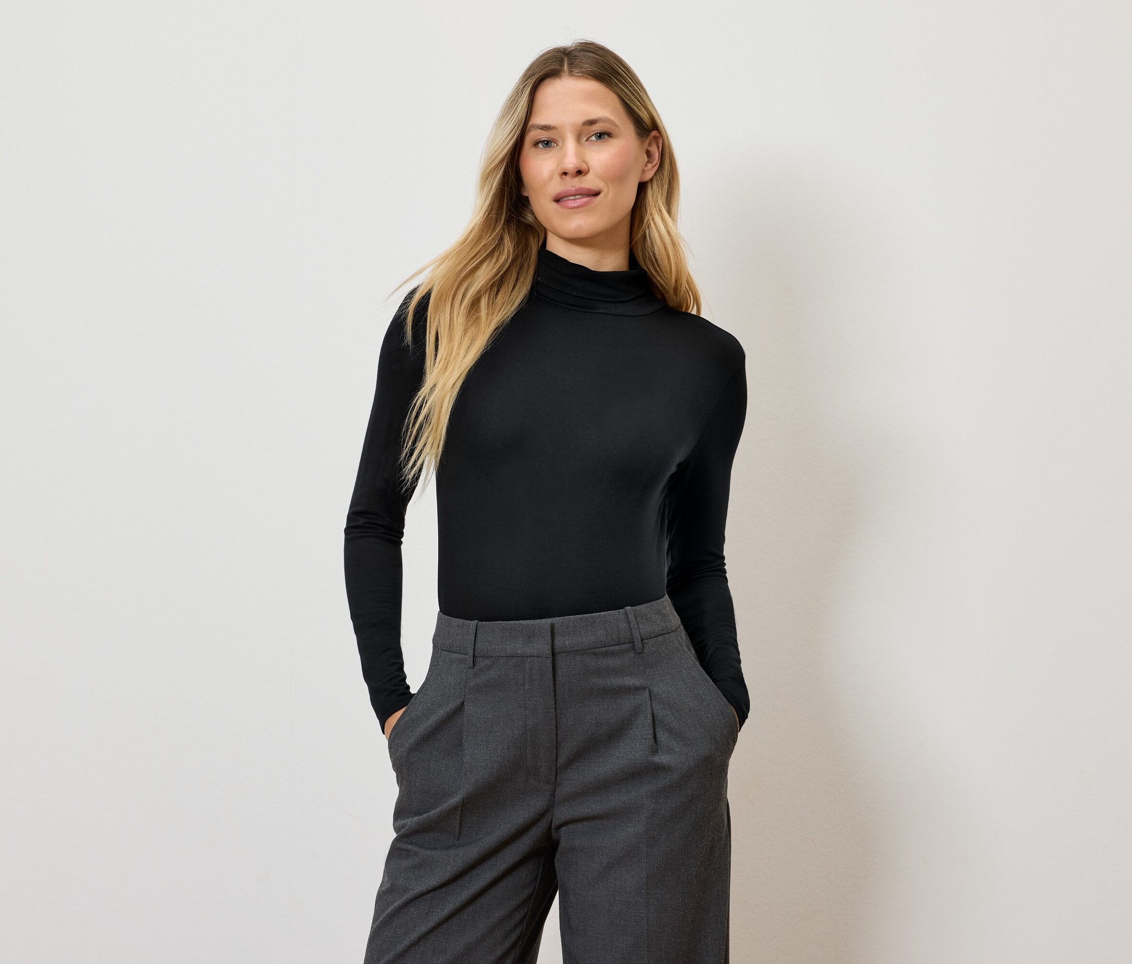 Uzun sarı saçlı bir kadın, siyah balıkçı yakalı kazak ve gri pantolonla poz veriyor.