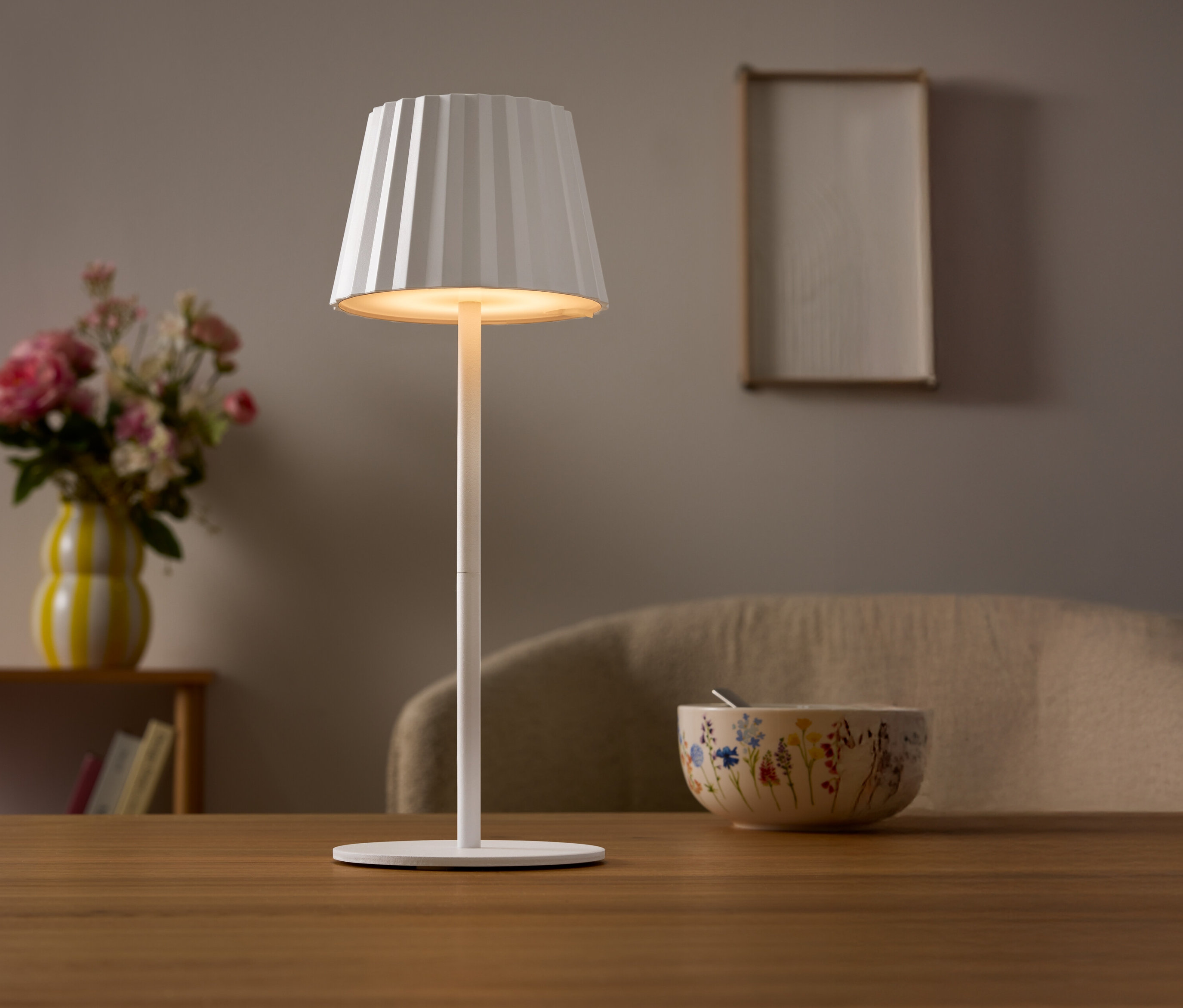 Masada pileli abajurlu beyaz bir lamba duruyor. Arka planda çiçek vazosu ve bir kase var.