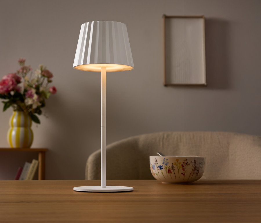 Masada pileli abajurlu beyaz bir lamba duruyor. Arka planda çiçek vazosu ve bir kase var.