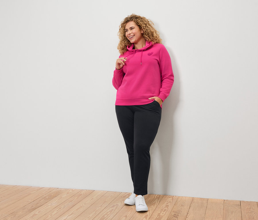 Kıvırcık saçlı bir kadın pembe kapüşonlu ve siyah pantolon giyiyor.