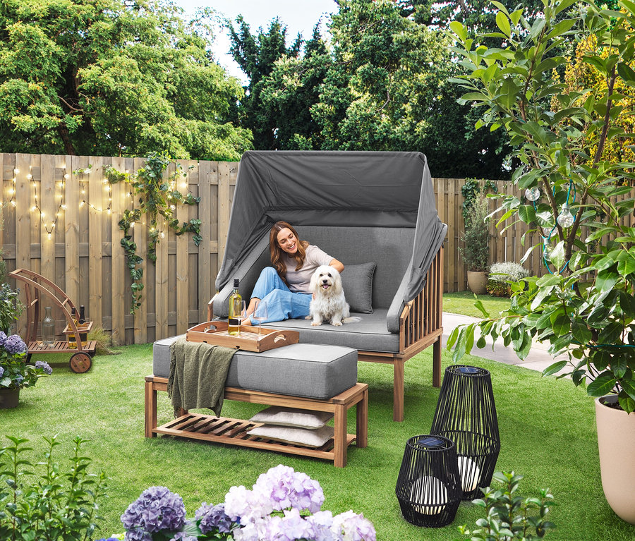 Bir kadın, Premium Lounge-Strandkorb mit Sonnenschutz-Dach'da bir köpekle oturuyor. Çimde Tekstil Bantlı Güneş Enerjili Fener, Büyük ve Tekstil Bantlı Güneş Enerjili Fener, Küçük duruyor. Arka planda çıkarılabilir tepsili bir bar arabası var.