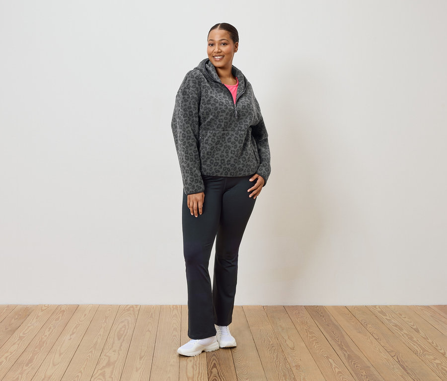 Kadın ayakta duruyor ve gri-siyah leopar desenli polar kapüşonlu, siyah pantolon ve beyaz spor ayakkabı giyiyor.