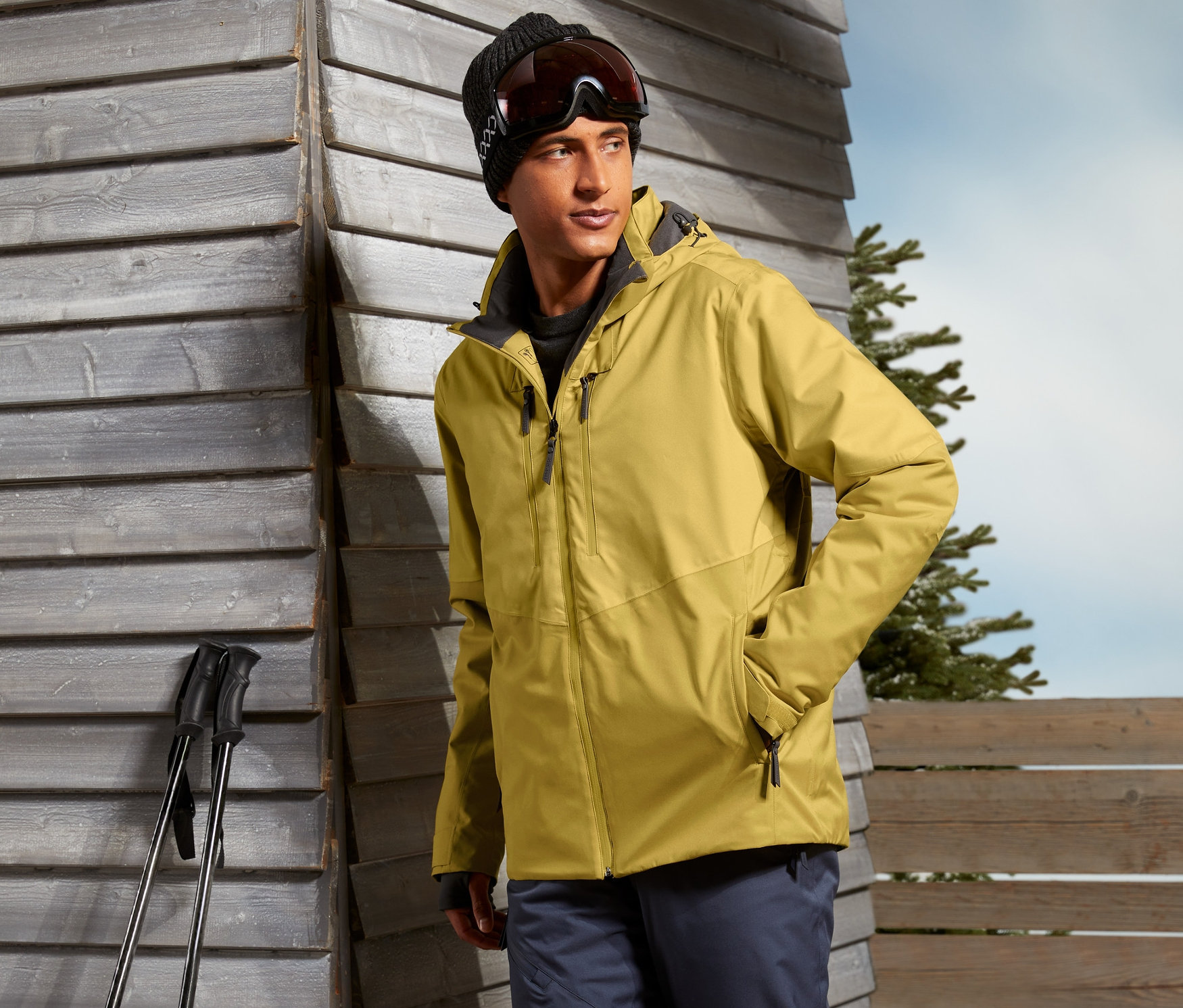 Hardal rengi kayak montu, kayak pantolonu ve botları giyen bir adam, kayak sopalarıyla dışarıda duruyor.